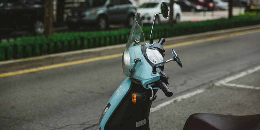 A blue-green moped is parked on a street, with a clear windshield, mirrors, and a headlight visible.