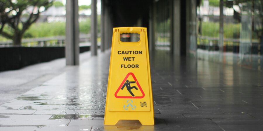 A yellow 'Caution Wet Floor' sign on a outdoor patio along the side of an office building with small puddles and other indications of a wet floor.