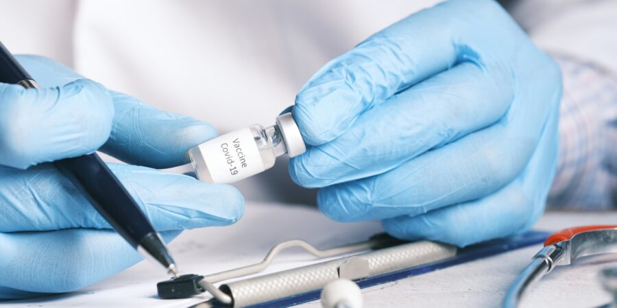 A medical professional looking closely at a vial of Covid-19 vaccine and a pen in their hand, indicating readiness to take notes on the clipboard from info they are reading from the vial.